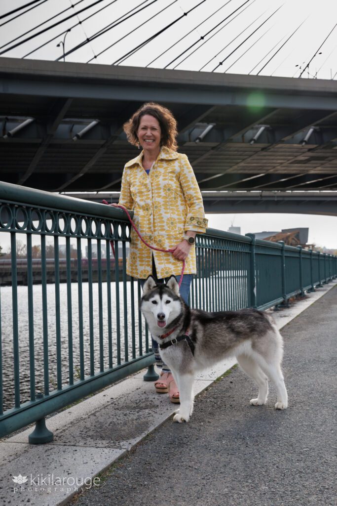 Woman smiling with sun flare under the Zakim Bridge Boston with her husky leaning on green railing next to the Charles River