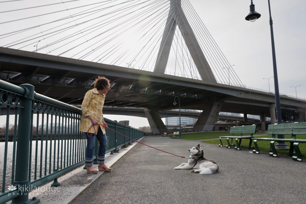 Woman in vintage yellow jacket and jeans laughing at her stubborn dog laying on the payment looking back at her under the Zakim Bridge in Boston MA