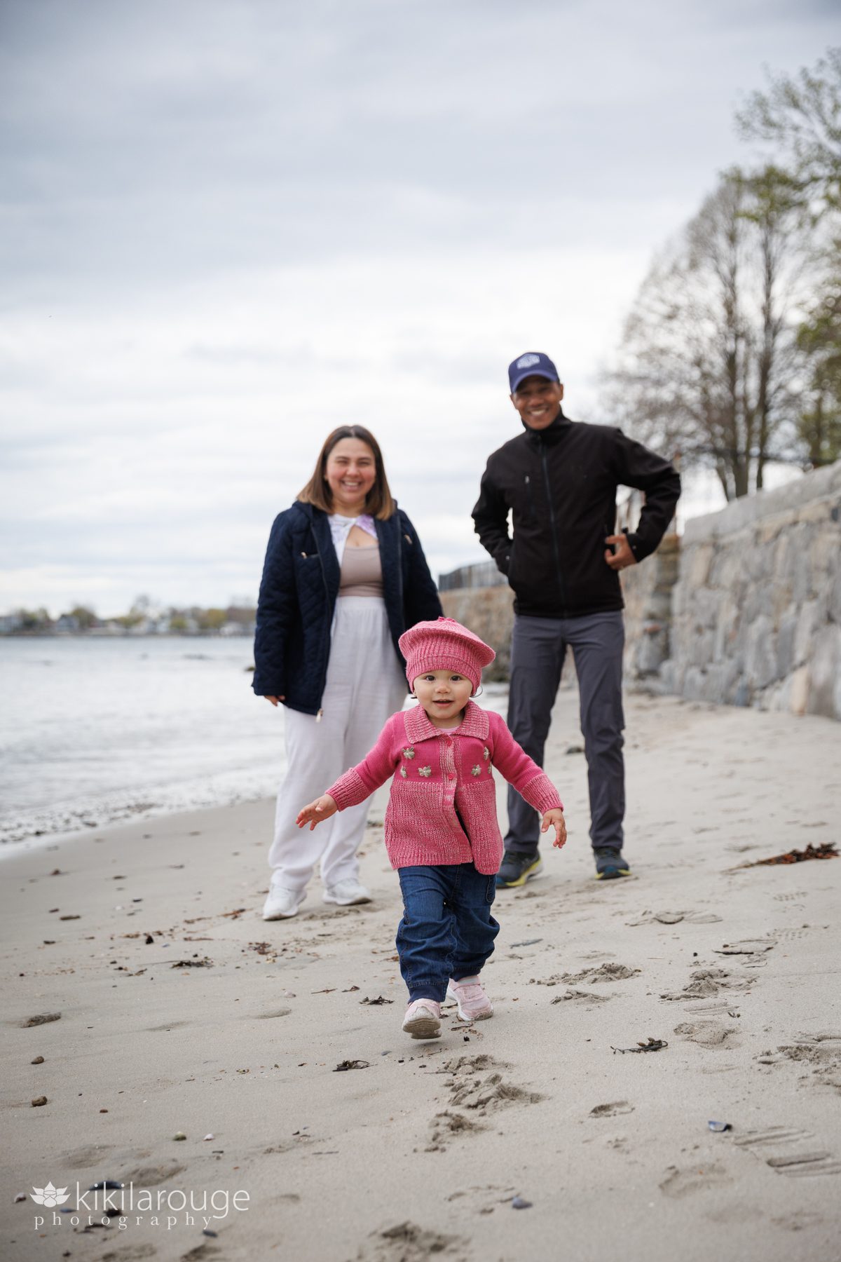 Little girl dressed in pink sweater and hat running towards the viewer with her doting parents looking on in the backdrop