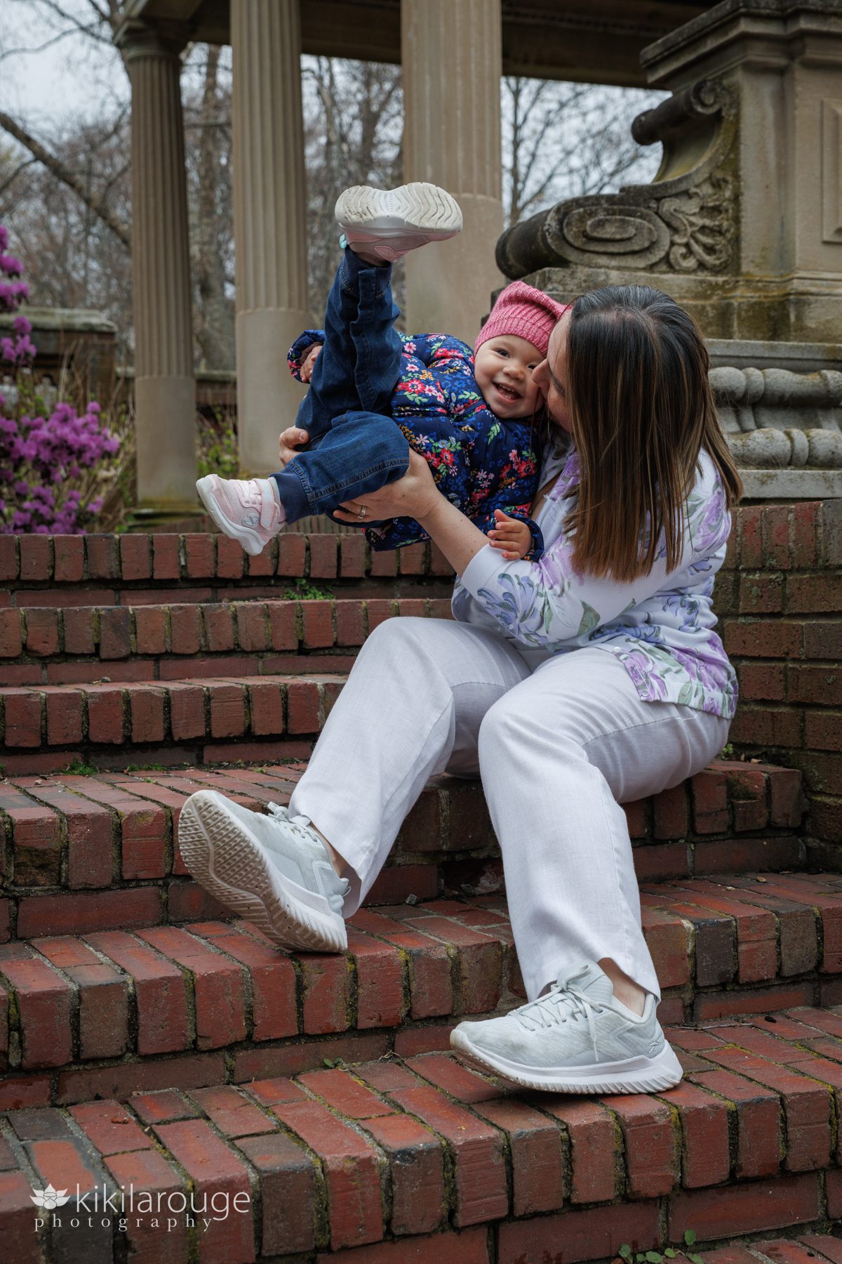 Mom in white linen and floral top sitting on brick steps tossing her daughter up in the air with little legs kicking and both smiling 