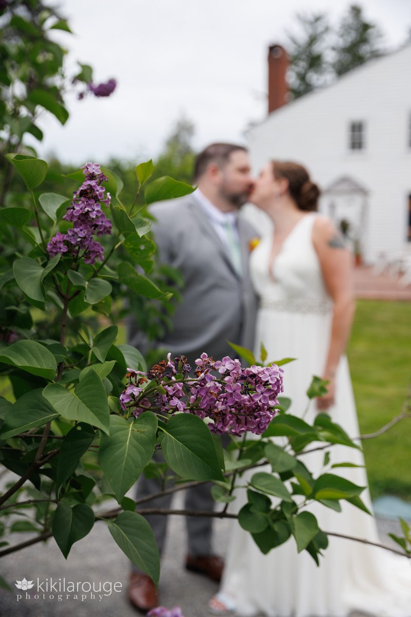 Purple lilacs in focus with couple in backdrop kissing and white Stephens Coolidge house in backgroun
