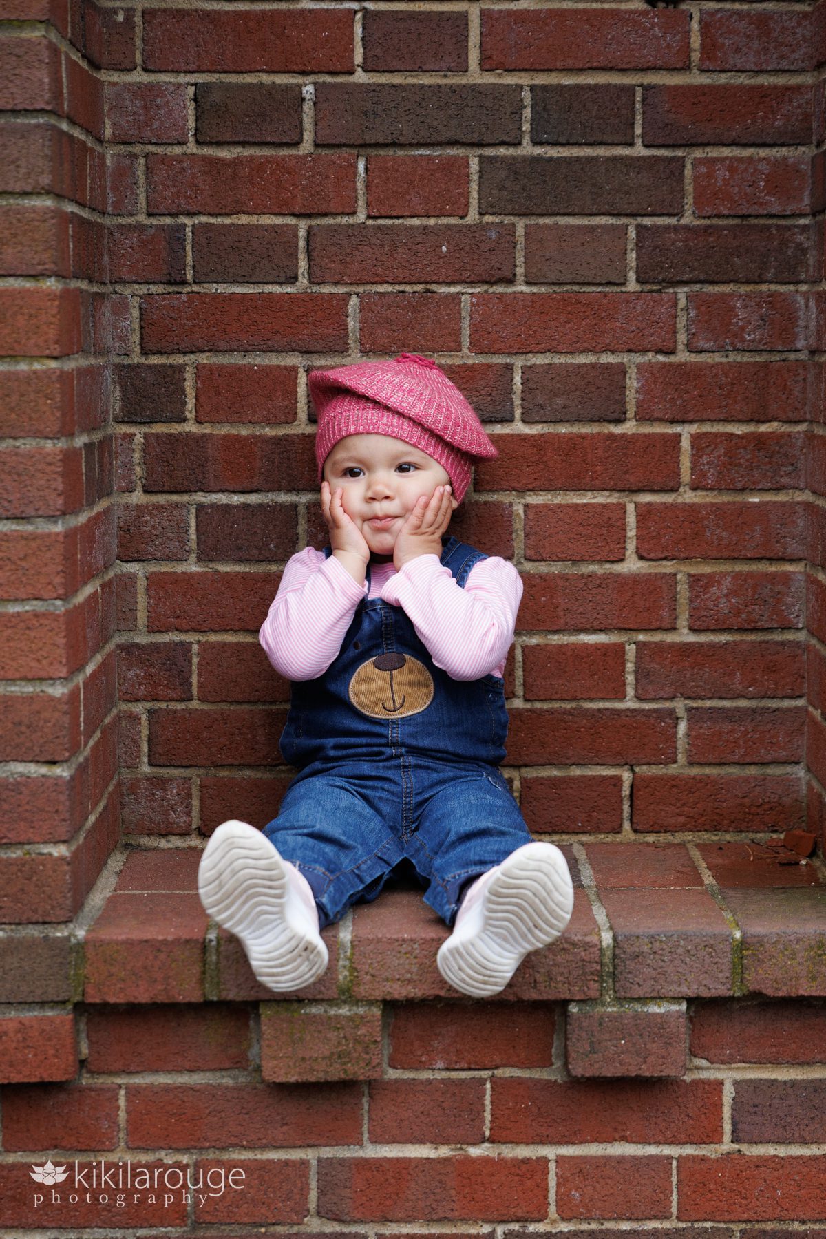 Little girl with her hands on her cheeks in denim overalls with a bear on the bib sitting in brick nook at Lynch Park rose garden