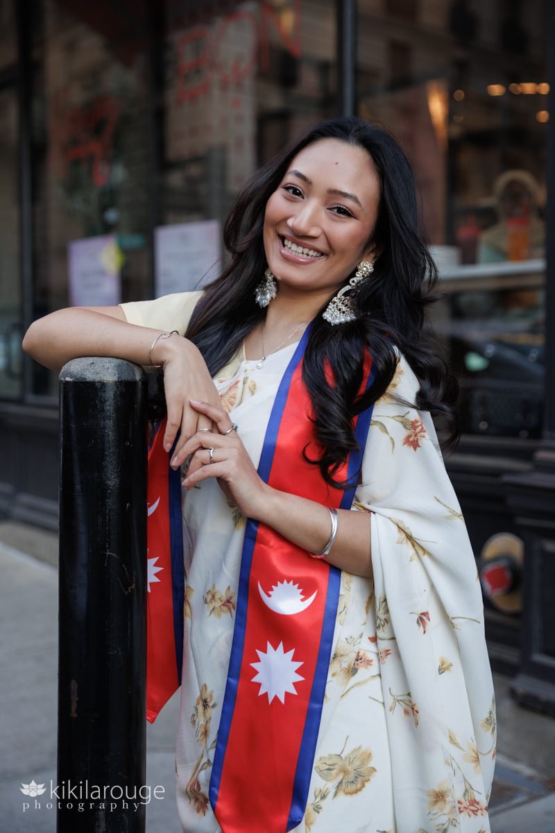 Beautiful Nepali graduate from Suffolk University leaning on black pole with pale yellow hair and Nepali graduation stole with a big smile