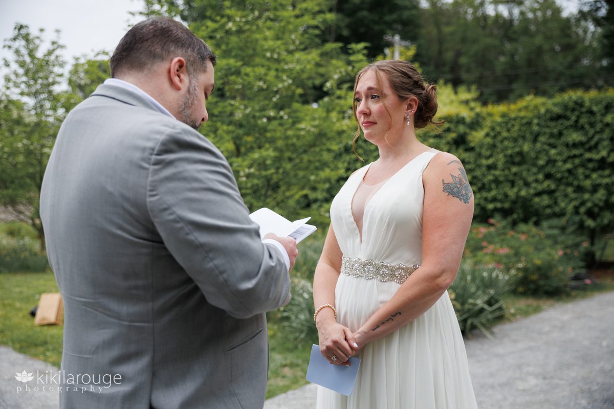 Groom in gray suit reading his wedding vows to bride who is teary eyed with emotion in beautiful lush green garden