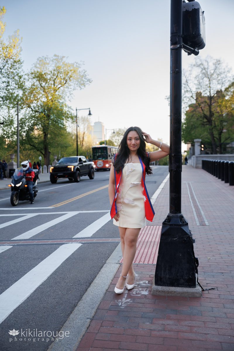 Woman in short pale yellow dress with 3D white flowers leaning on a traffic light in front of the MA State House with motorcycle, Boston Trolley and the Prudential building in the backdrop