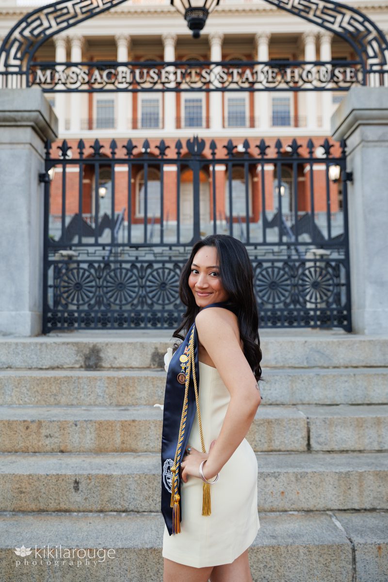 Woman with long black hair and light yellow dress and graduation stole and tassels in front of the MA State House looking over her shoulder