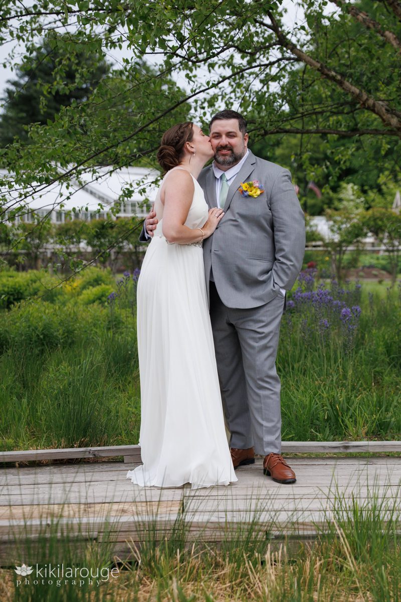 Wedding couple on wood boardwalk at Trustees garden bride kissing the groom in gray suit on cheek