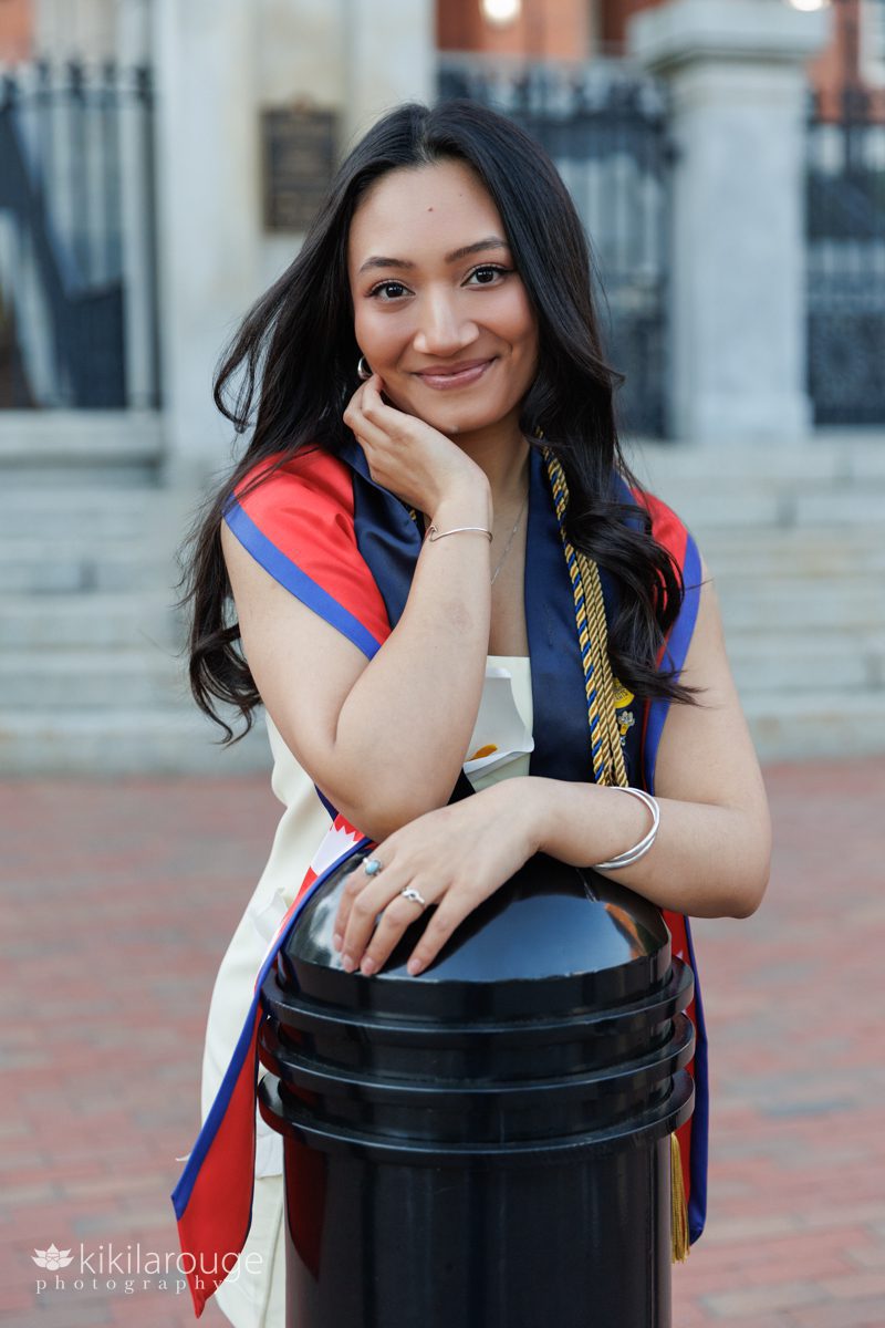 Pretty Nepali woman with graduation stoles and tassels leaning on black barrier post at MA State House