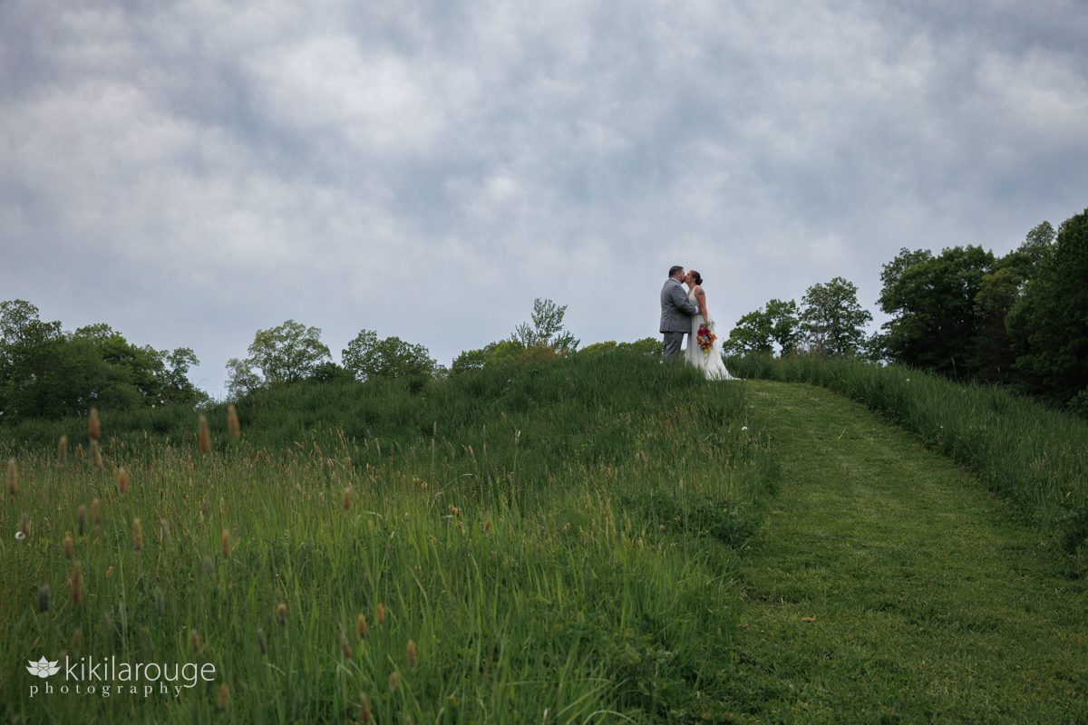 Wedding couple kissing atop a hill in a large meadow with dramatic overcast skies