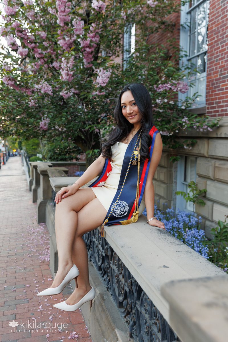 Woman sitting on concrete fence with Spring blooms all around on Beacon Street Boston wearing a short yellow floral dress and her graduation stoles and pins