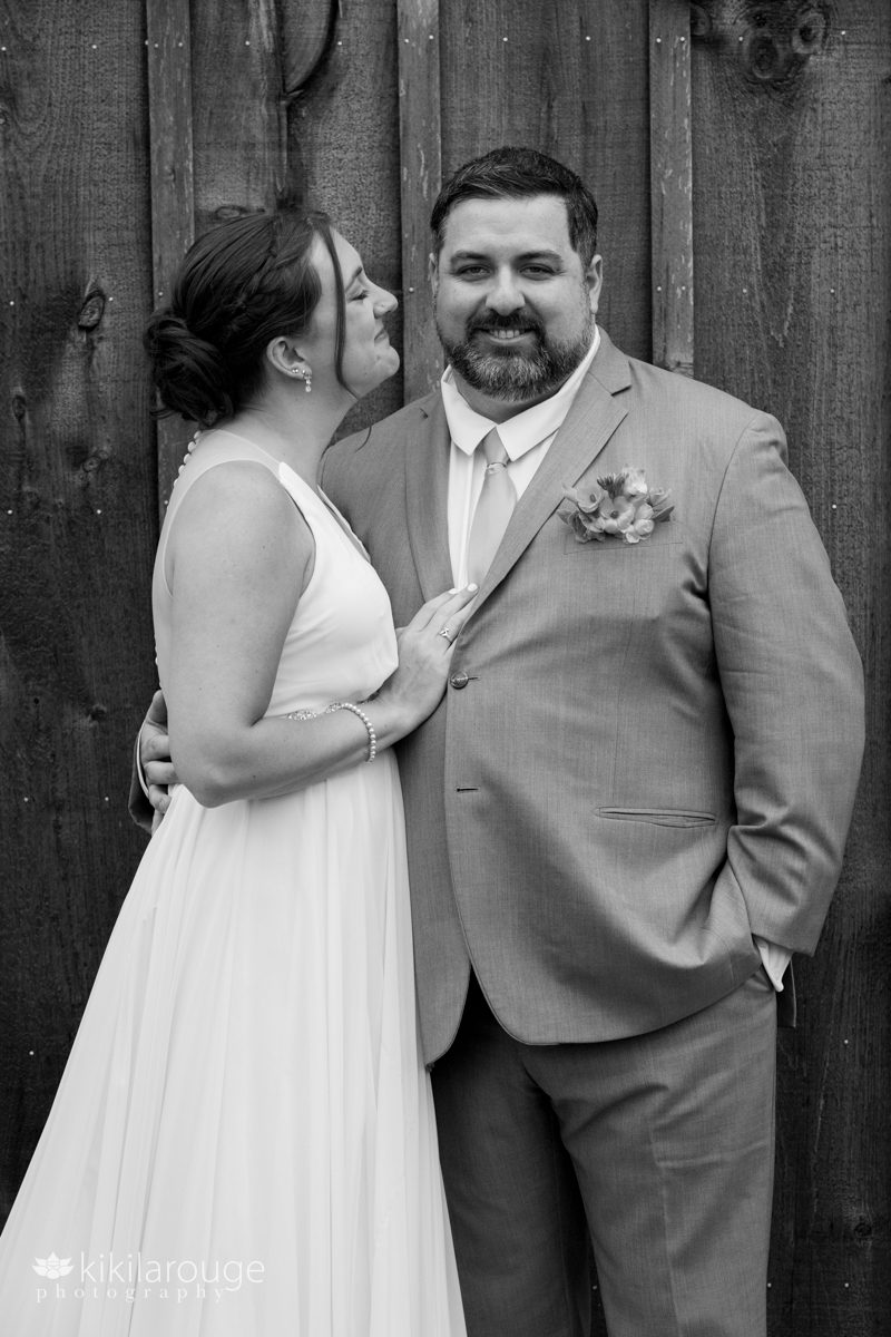 BW of a couple next to gray wood building bride looking and smiling at groom