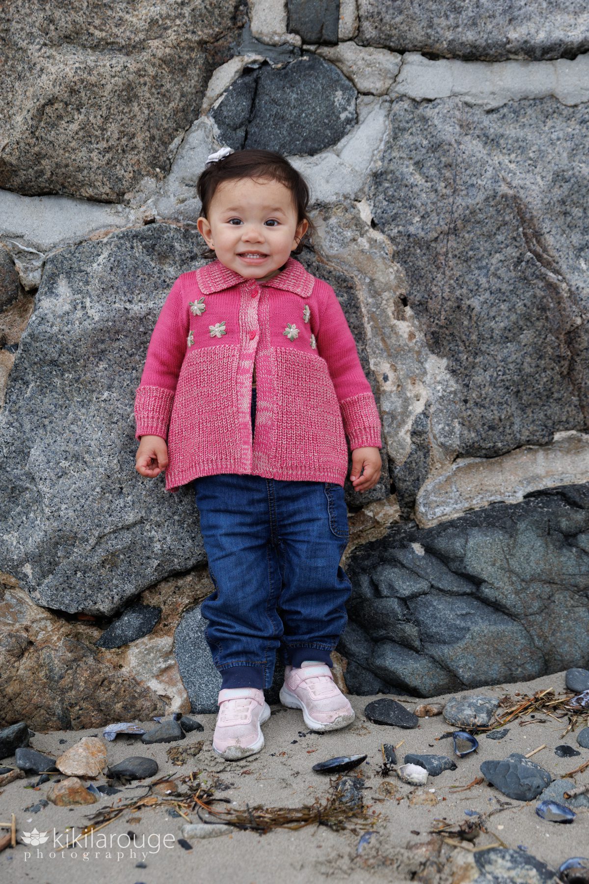 Cute little 1.5 year old girl leaning against a stone wall at the beach smiling with white bow in her hair and pink sweater with flowers and denim jeans
