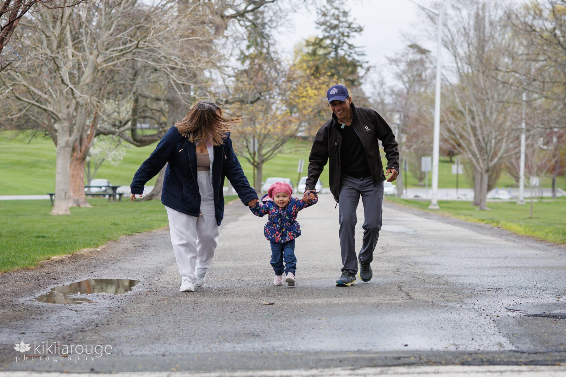 Couple running down paved road in park with toddler in a pink beret hat and floral winter coat