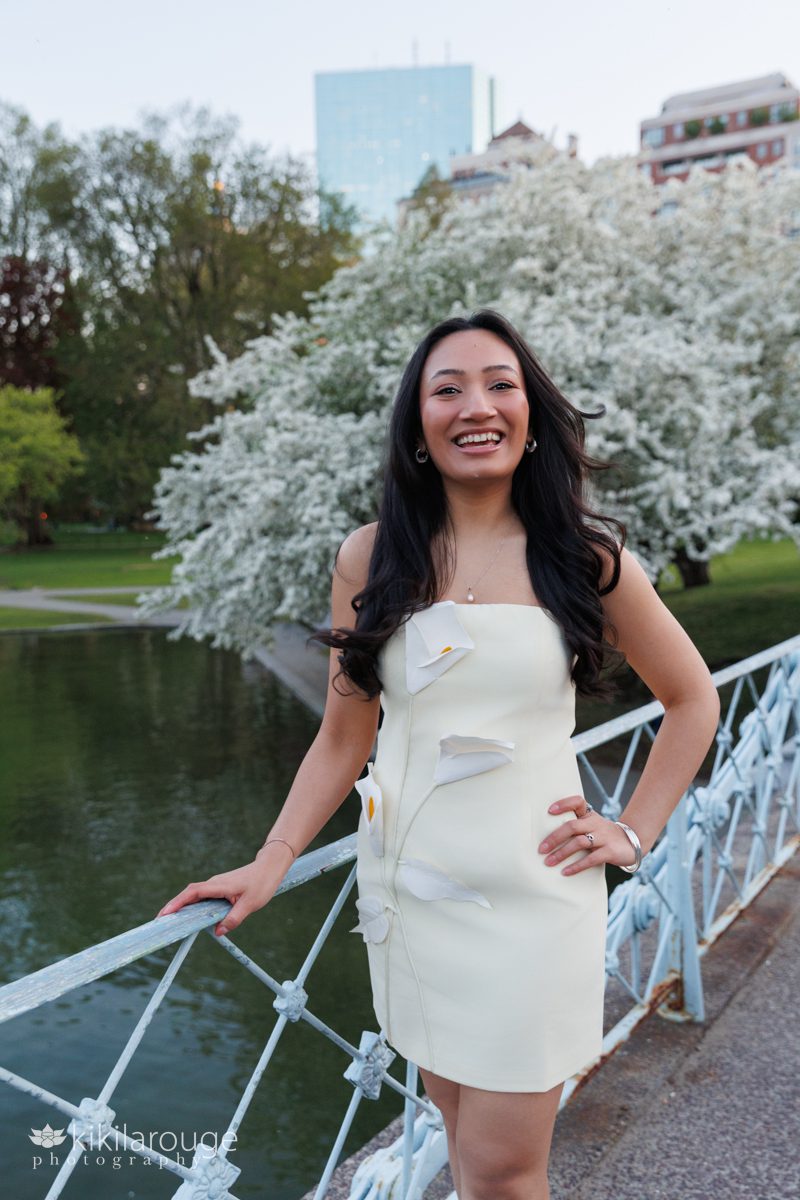 Woman with long black hair smiling leaning on light blue metal bridge in Boston's Public Garden with the a huge tree in white bloom and the Pur in the backdrop