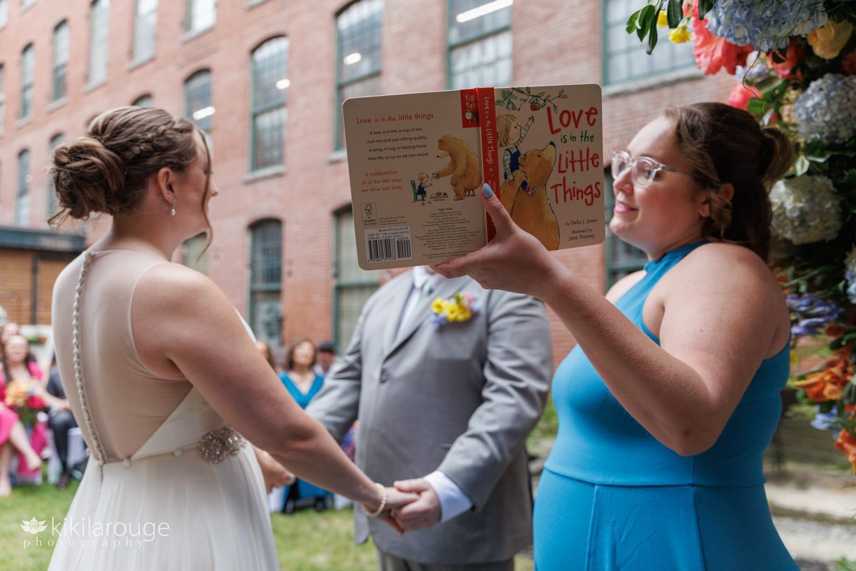 Woman holding up a book Love is in the Little Things reading it to a wedding couple at the altar on the wedding day in brick mill garden