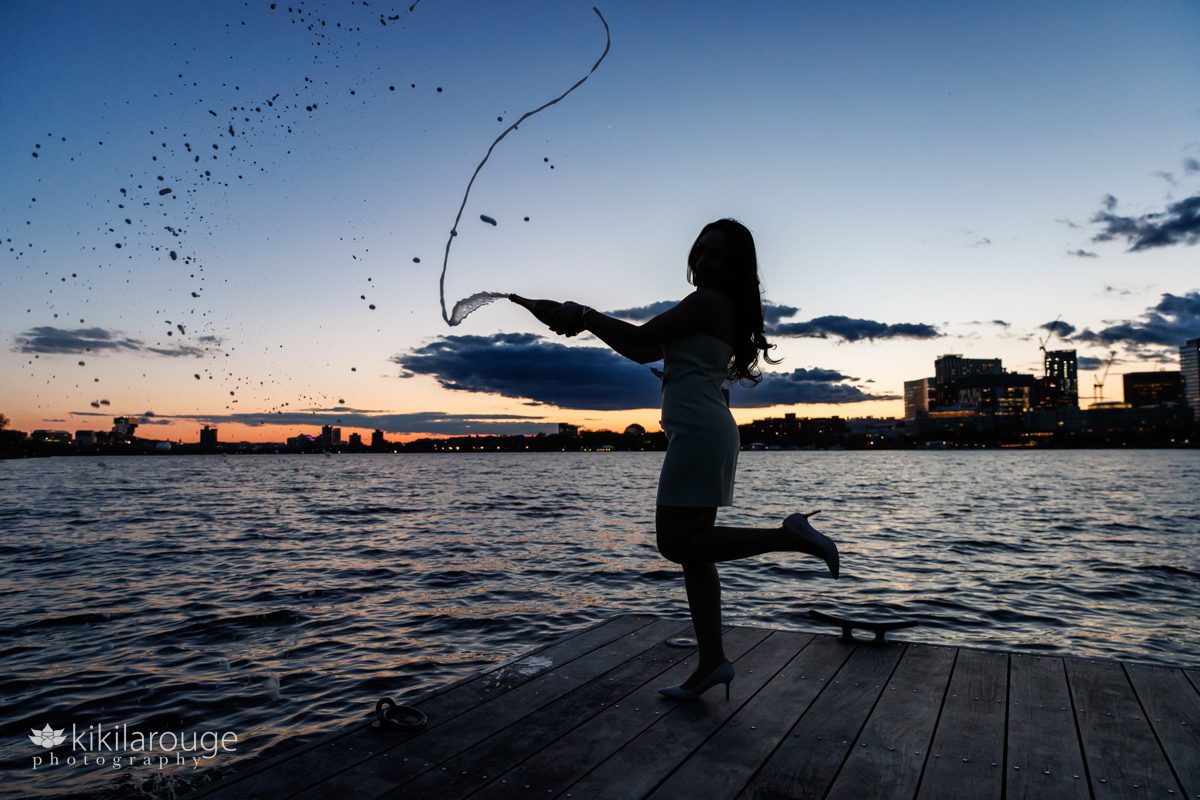 Silhouette on the Charles River dock at sunset with a woman kicking up her heal and spraying a bottle of champagne celebrating her college graduation