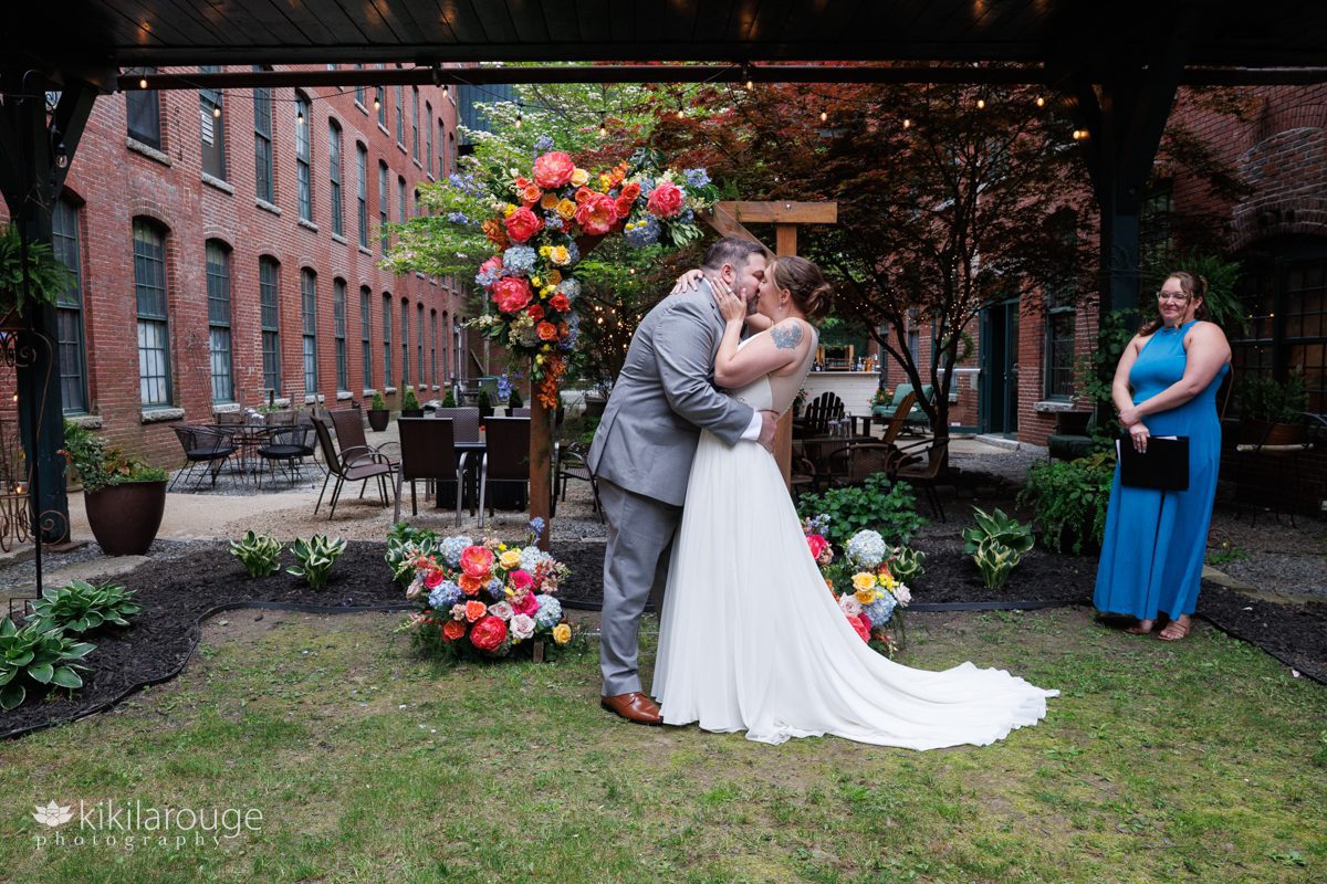 First kiss as married couple in front of wood altar adorned with colorful floral arrangements inside a brick wall courtyard