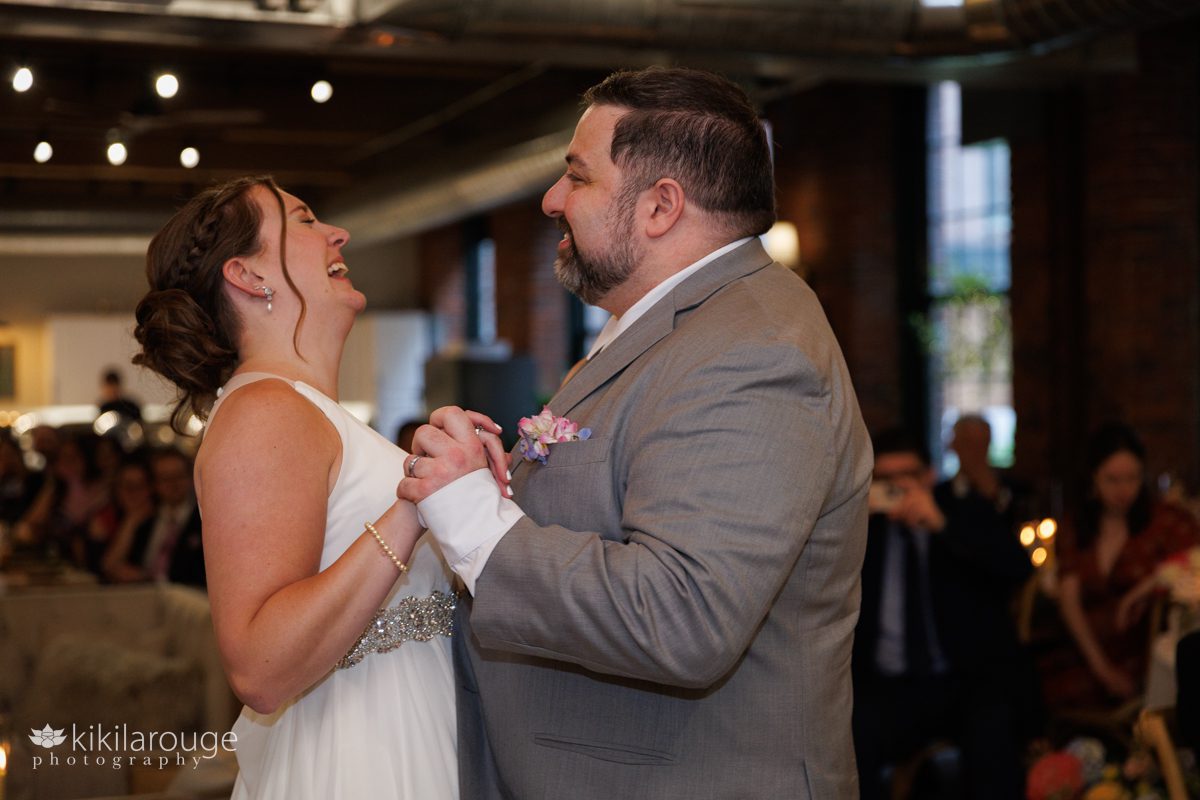 Couple's first dance with bride laughing and groom smiling at her and guest and family in the backdrop