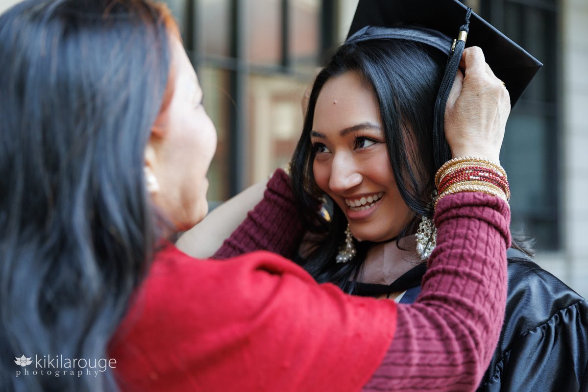 Mom helping her daughter put on her daughter's graduation cap with both smiling at each other 