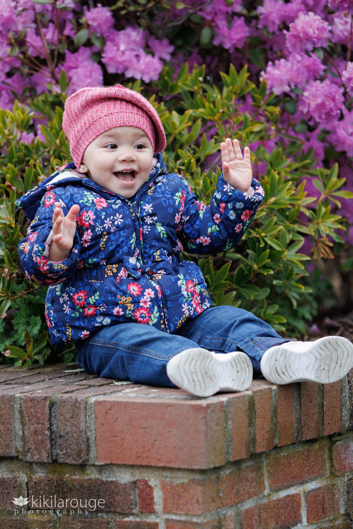 Toddler girl laughing with hands in air wearing pink beret like hat and a puffy floral navy coat.