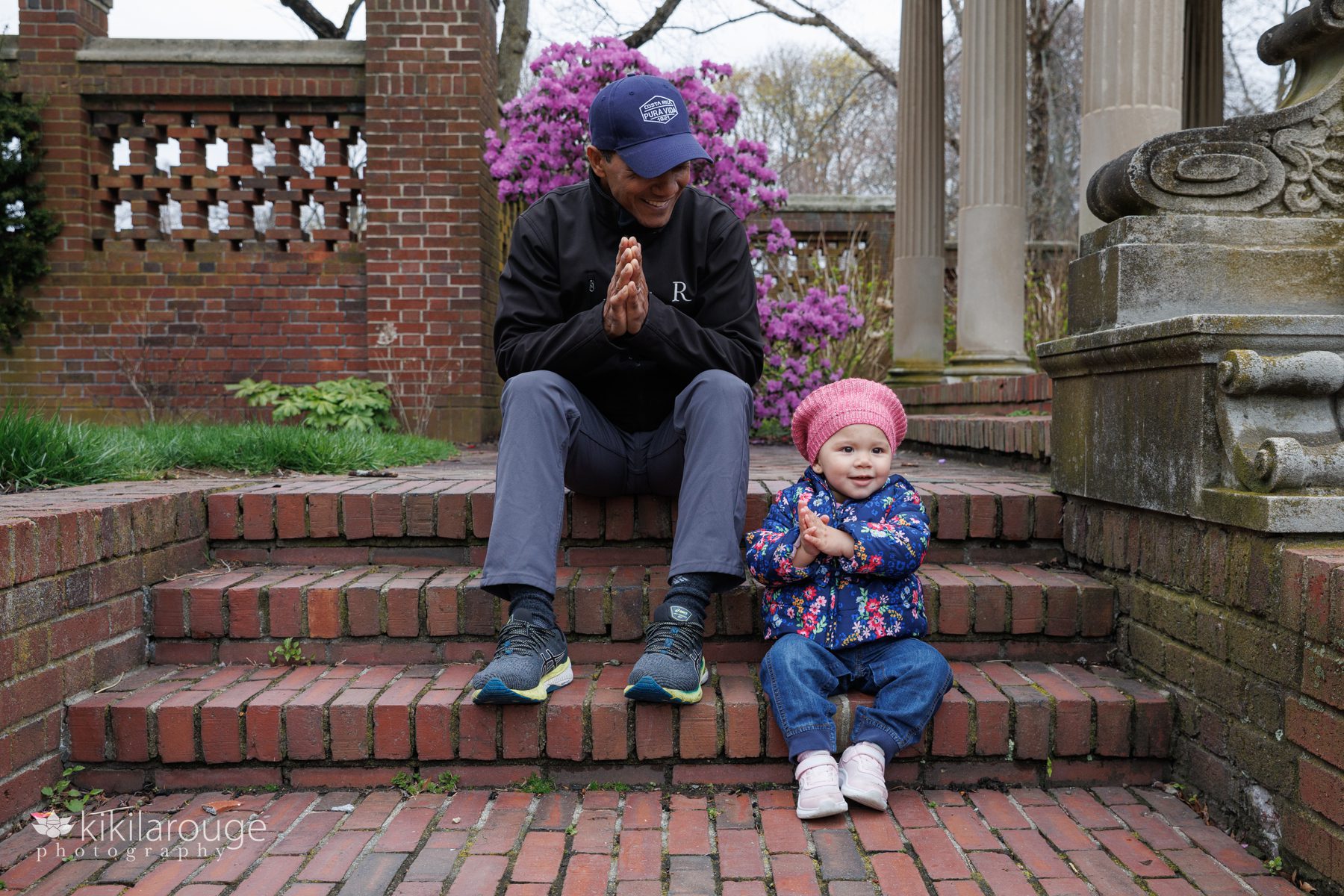 Man holding hands in Namaste pose smiling sitting on brick stairs looking down at his toddler daughter doing the same in a pink hat and floral coat