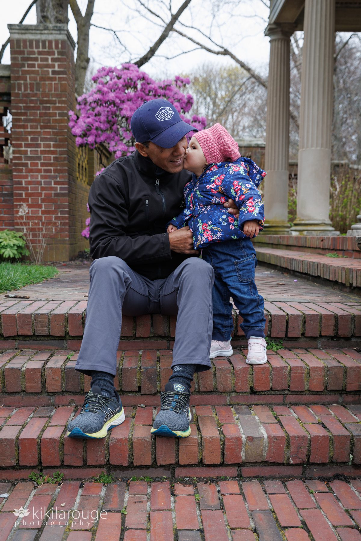 Little toddle girl in pink hat sitting on brick steps in Lynch Park rose garden giving her Dad a kiss on the cheek.