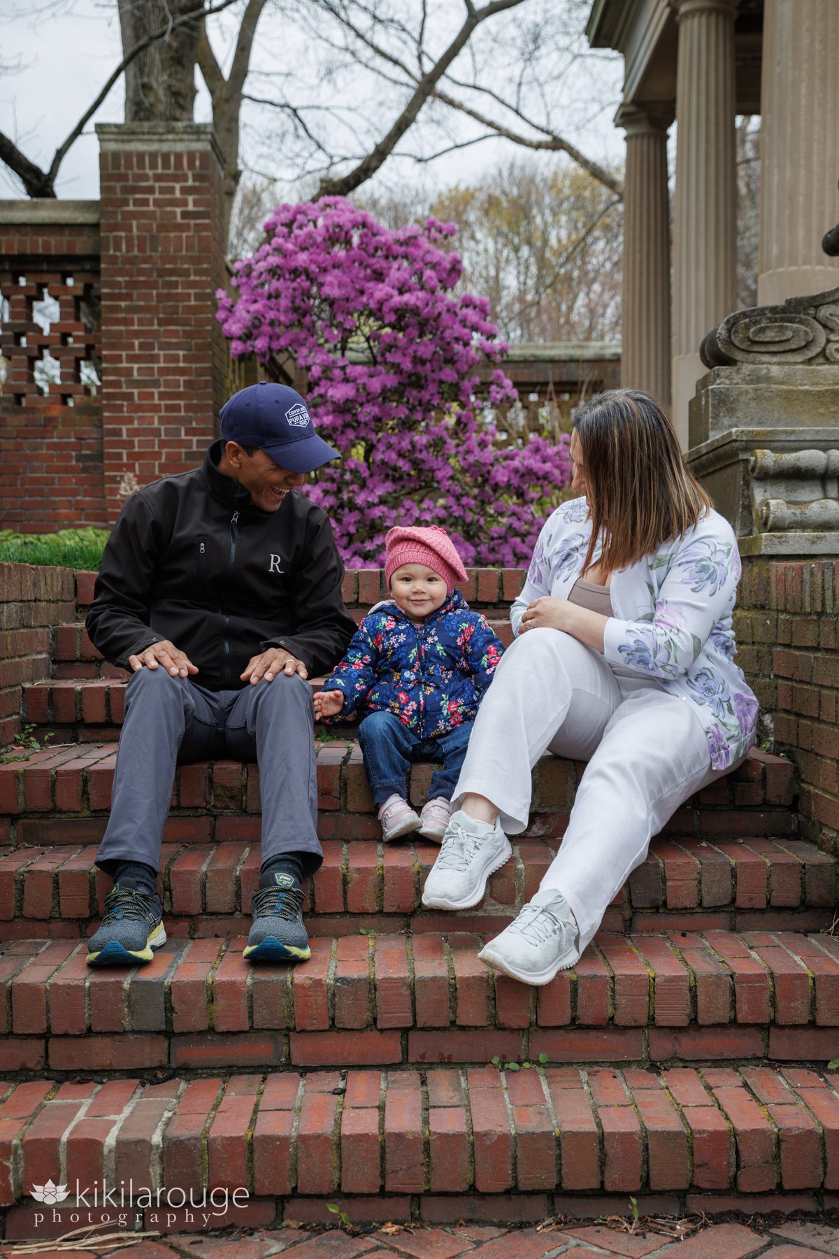 Couple sitting on brick steps both looking at and smiling at their cute toddler daughter in a pink hat and warm floral coat