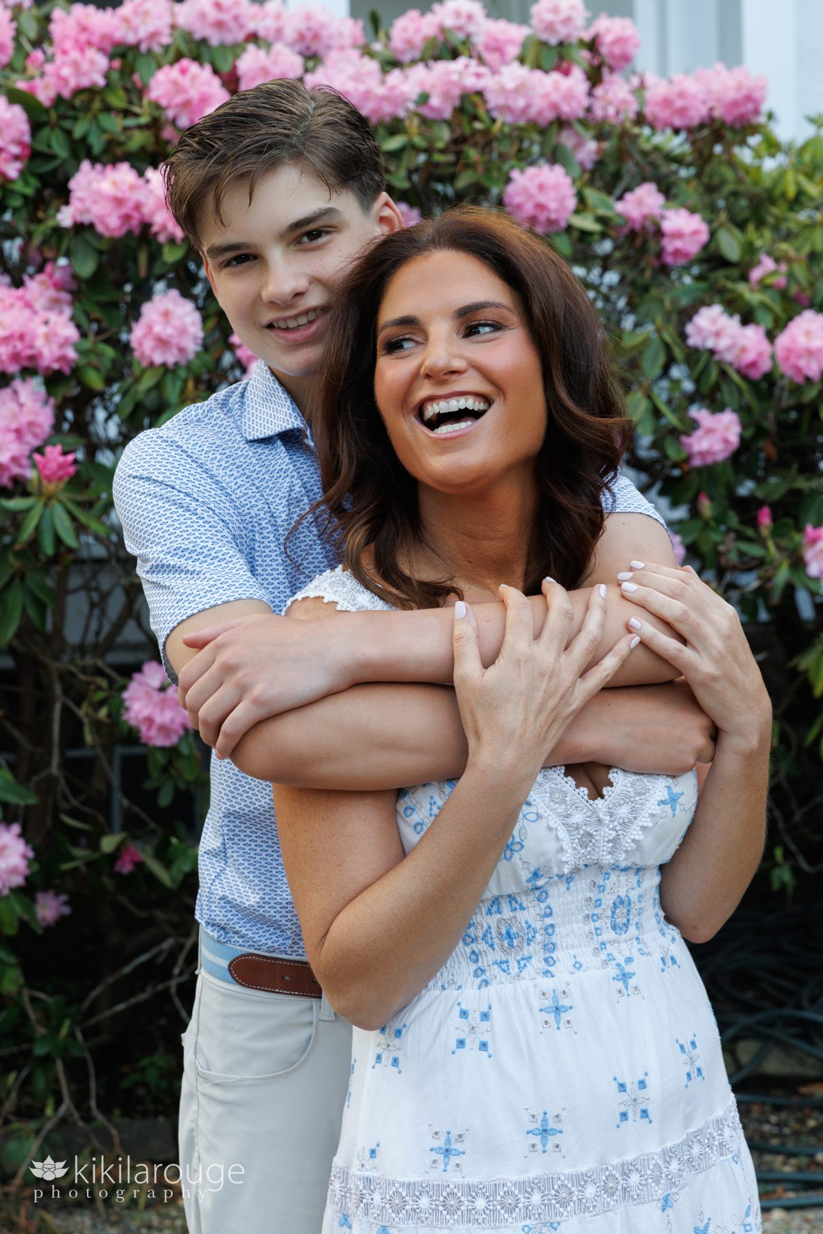 Son in light blue polo shirt hugging Mom who is laughing to the side in a white and blue sundress.
