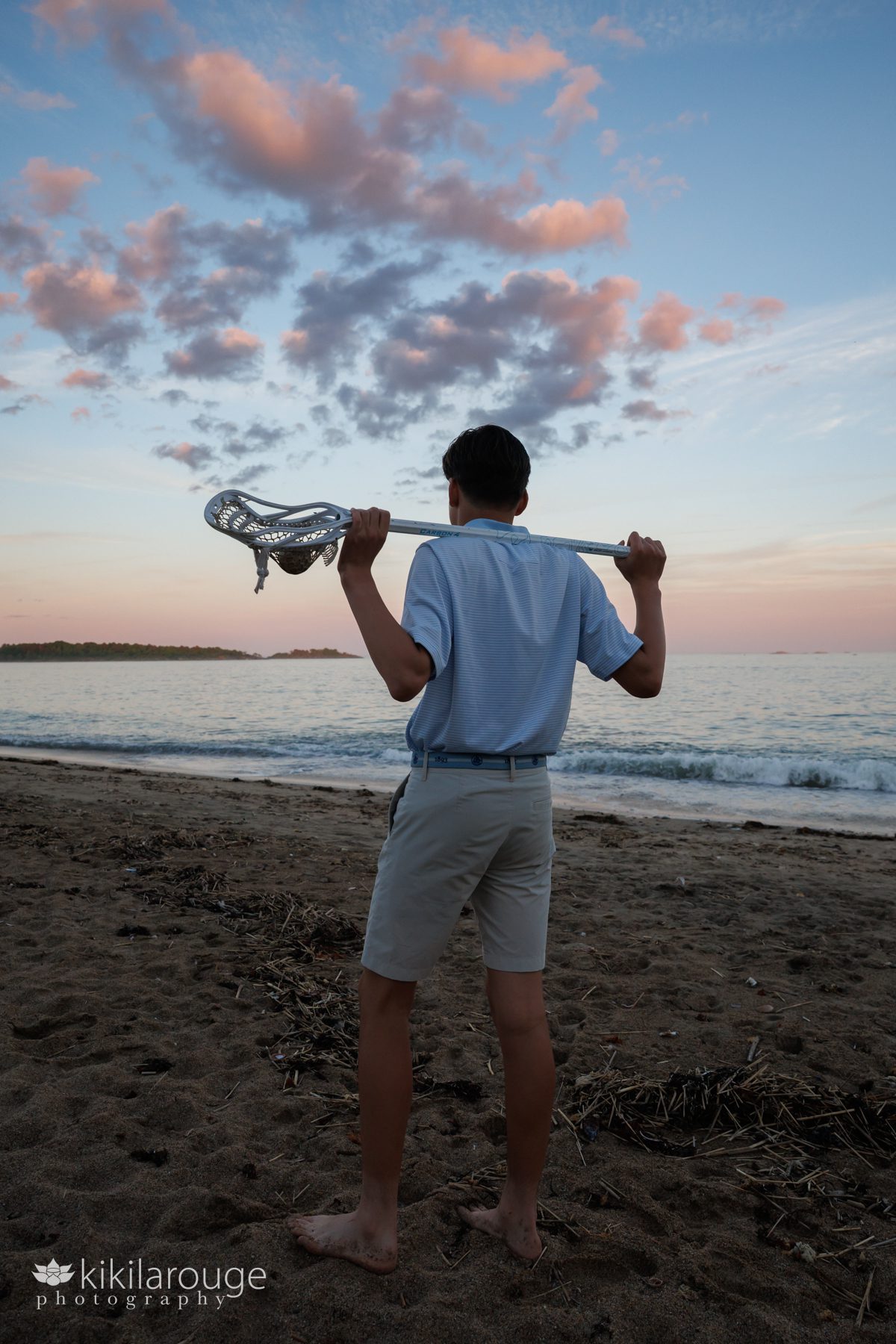 Boy holding a lacrosse stick over his shoulders looking out at pink sunset at the beach in silhouette 