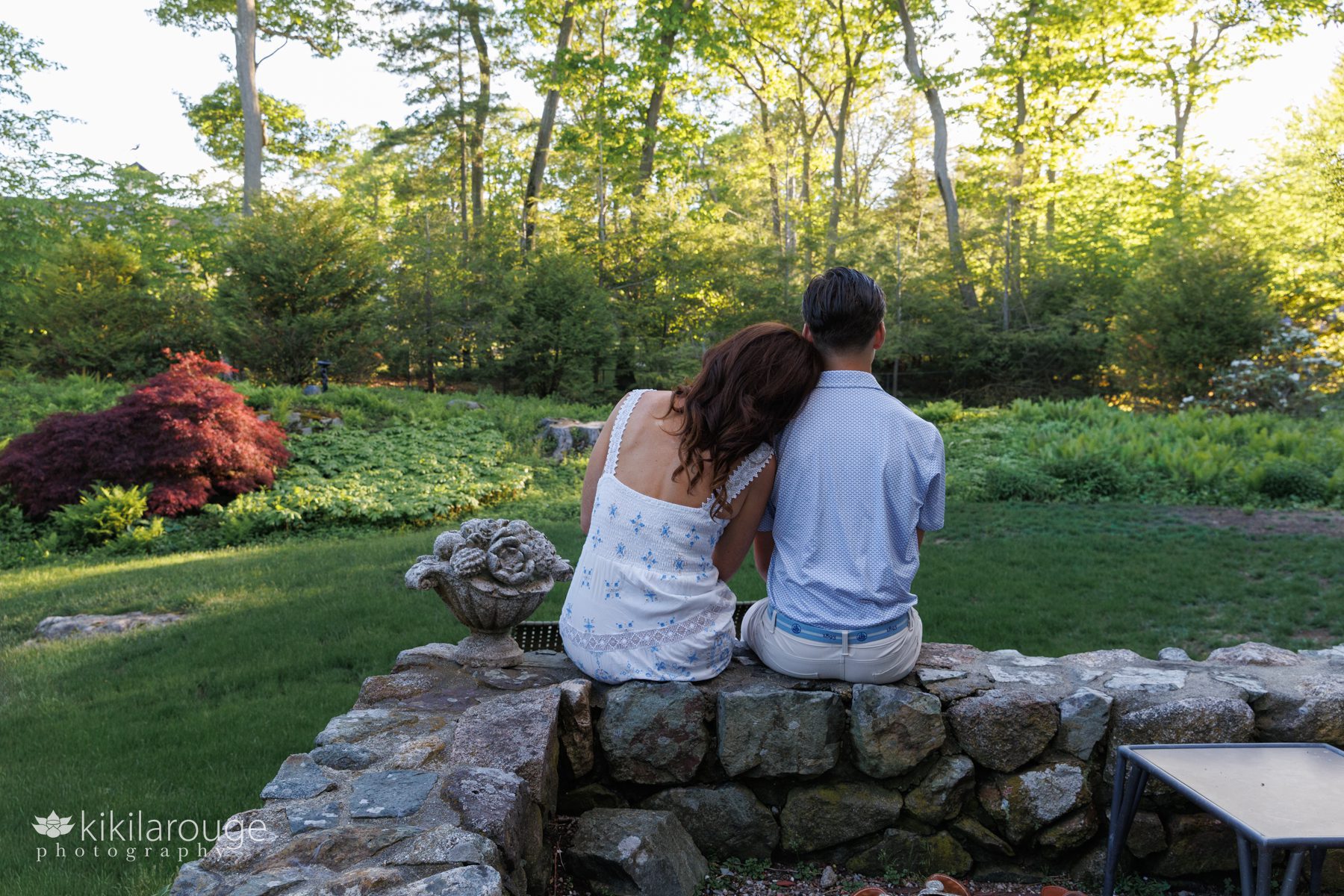 Mom in white sundress leaning on her son sitting on a stone wall looking out on their yard