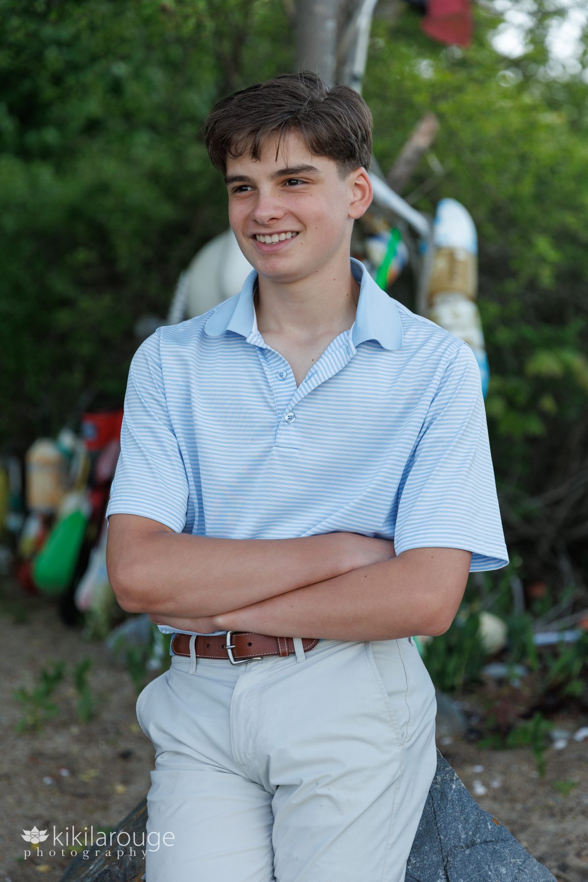 Young boy with stripped light blue shirt and arms crossed leaning on rock with buoys in the backdrop on beach path