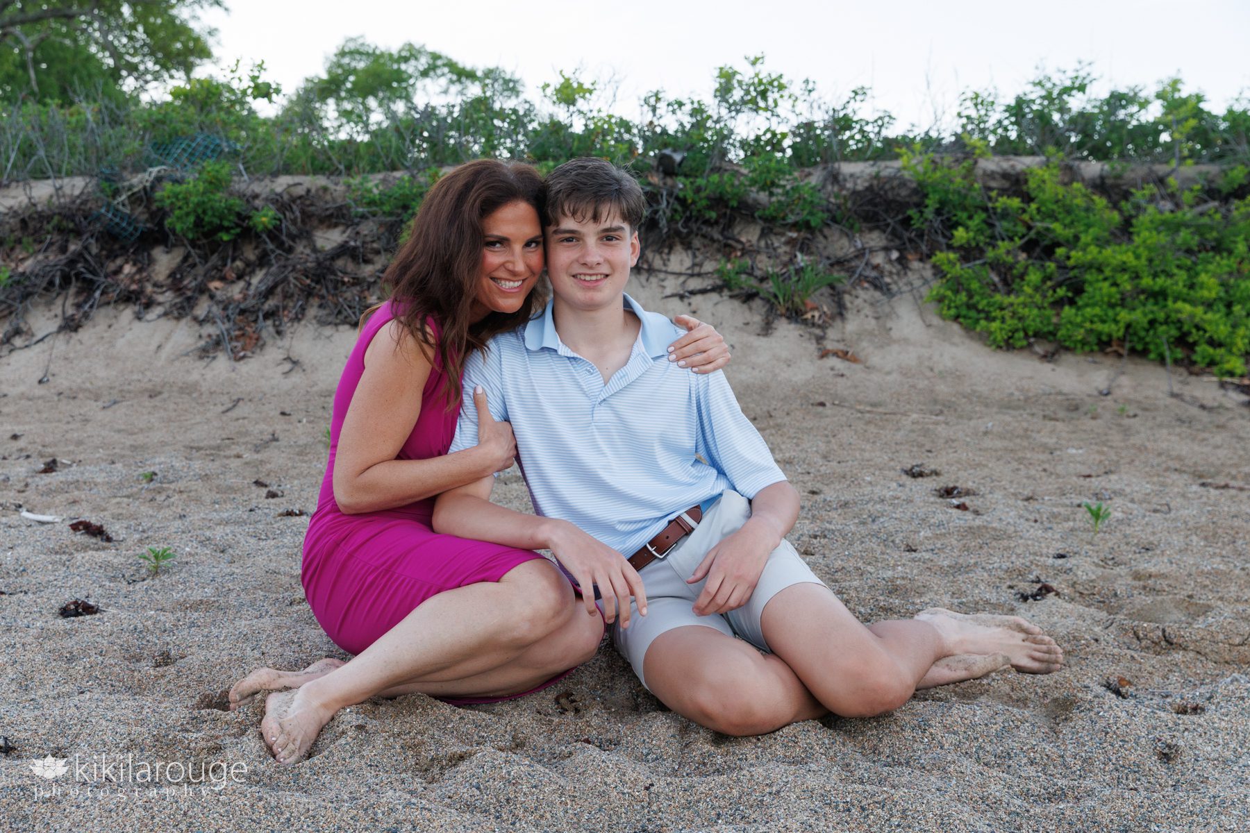 Mom in pink dress hugging her son sitting on the beach