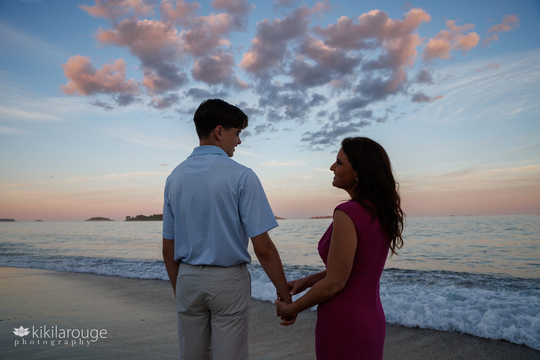 Silhouette of Mom in pink dress looking and smiling at her tall son at the beach with dramatic sunset of pink clouds