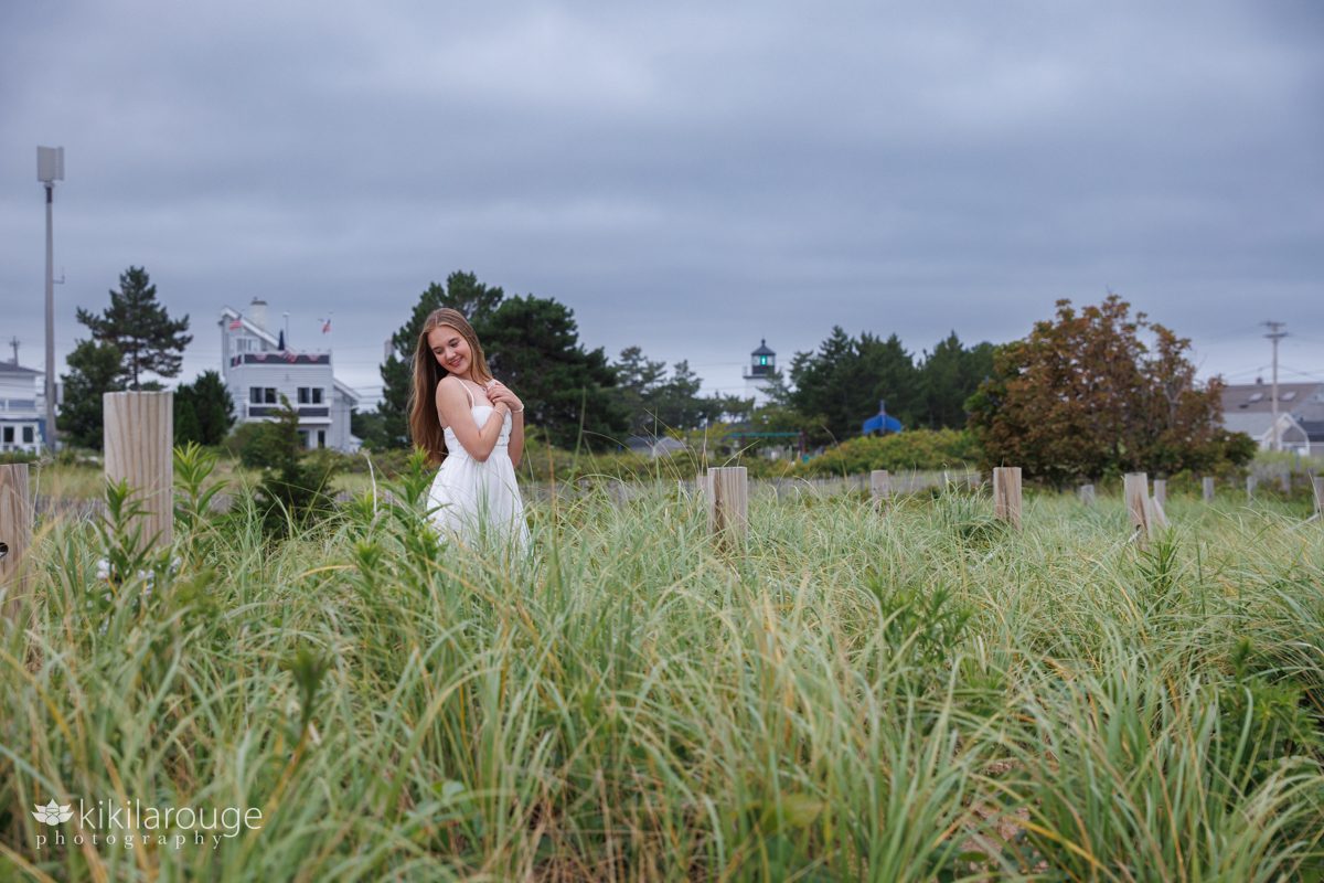 Girl smiling looking down standing in blowing dunes with Plum Island lighthouse in the background 