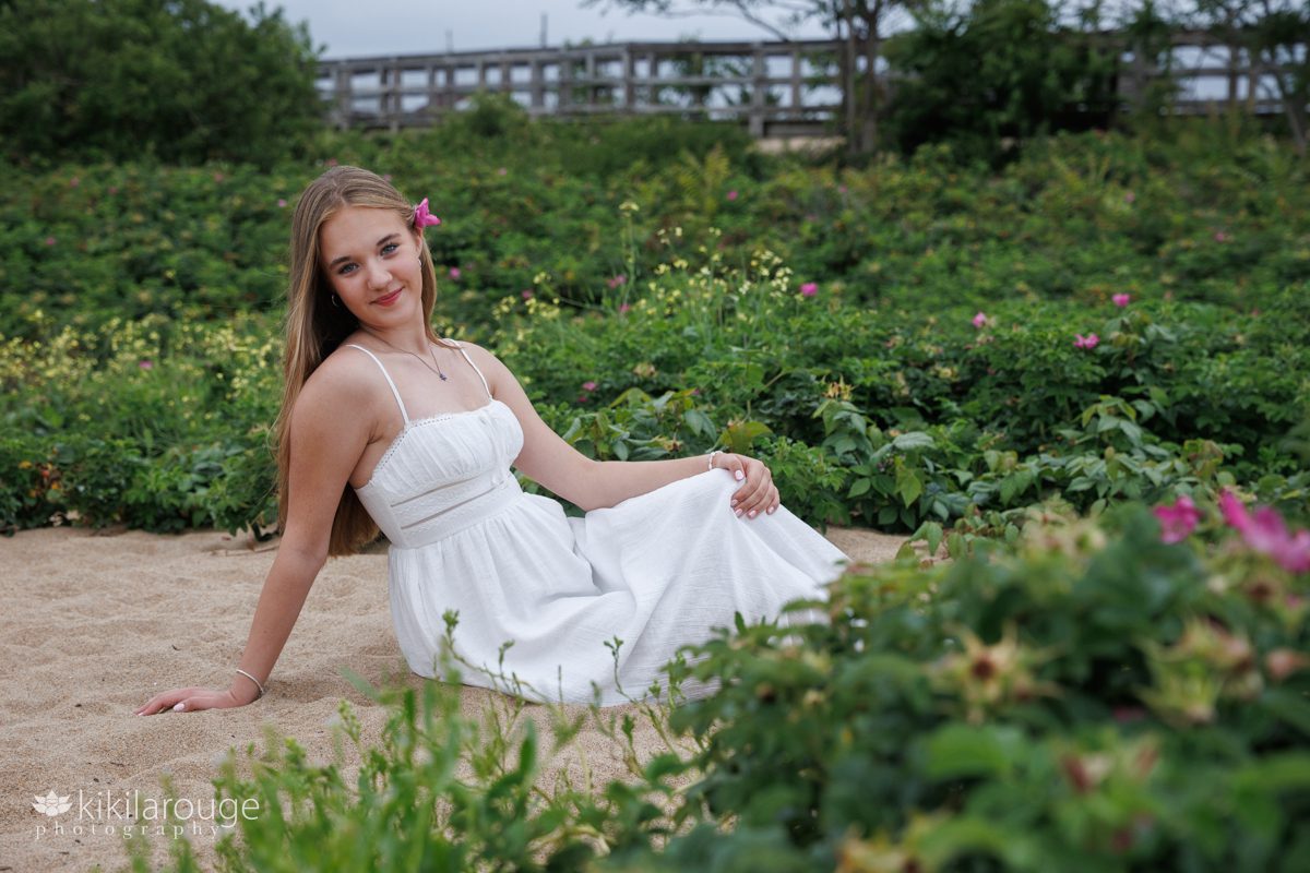 Senior portrait teen girl sitting on sand in white summer dress surrounded by beach rose bushes