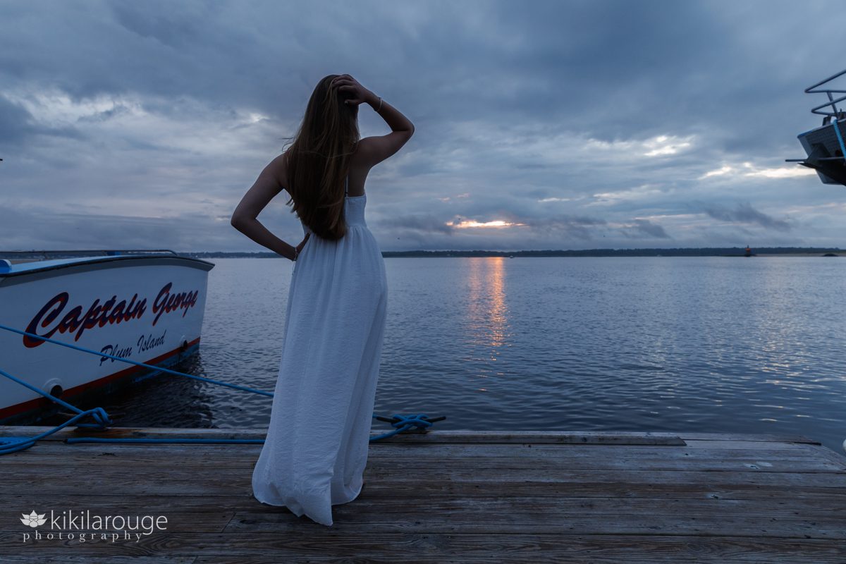 Silhouette of a teen girl with hand in her hair and dramatic skies with a peek of an sunset in clouds and reflecting on the water