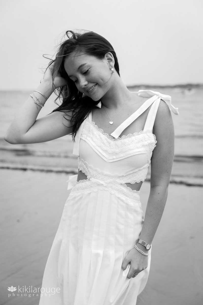 BW teen girl at beach water's edge in a Lacey sundress with wind blowing her hai as she looks down and smiles
