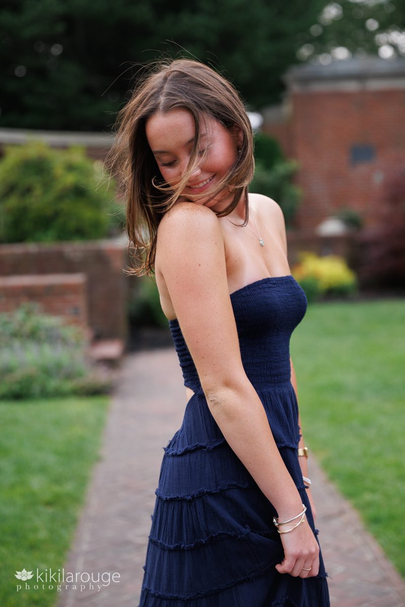 Teen senior girl in brick garden smiling looking down with hair blowing holding her long navy blue summer dress