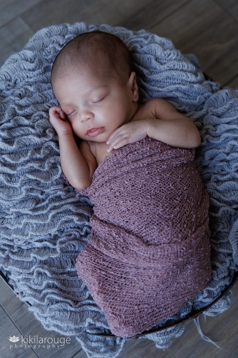 Newborn baby wrapped in mauve cheesecloth sleeping with one hand up by her face and other resting on her chest in a basket with a gray blanket