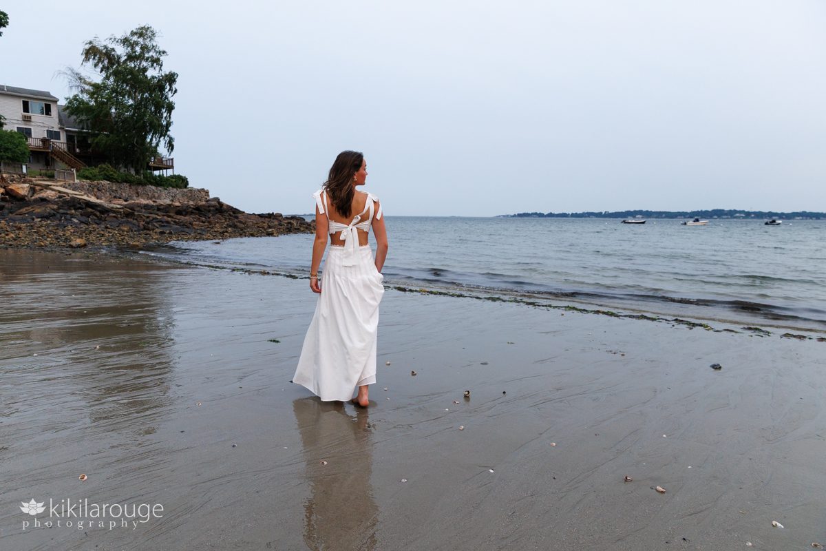 Senior teen girl in beautiful white dress with bows walking on beach at sunset with a reflection in the water as she looks to the side