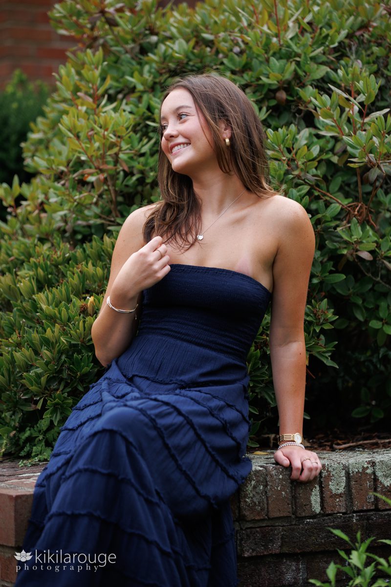 Teen girl Senior portrait sitting on brick wall in garden smiling off to the left in a navy summer dress