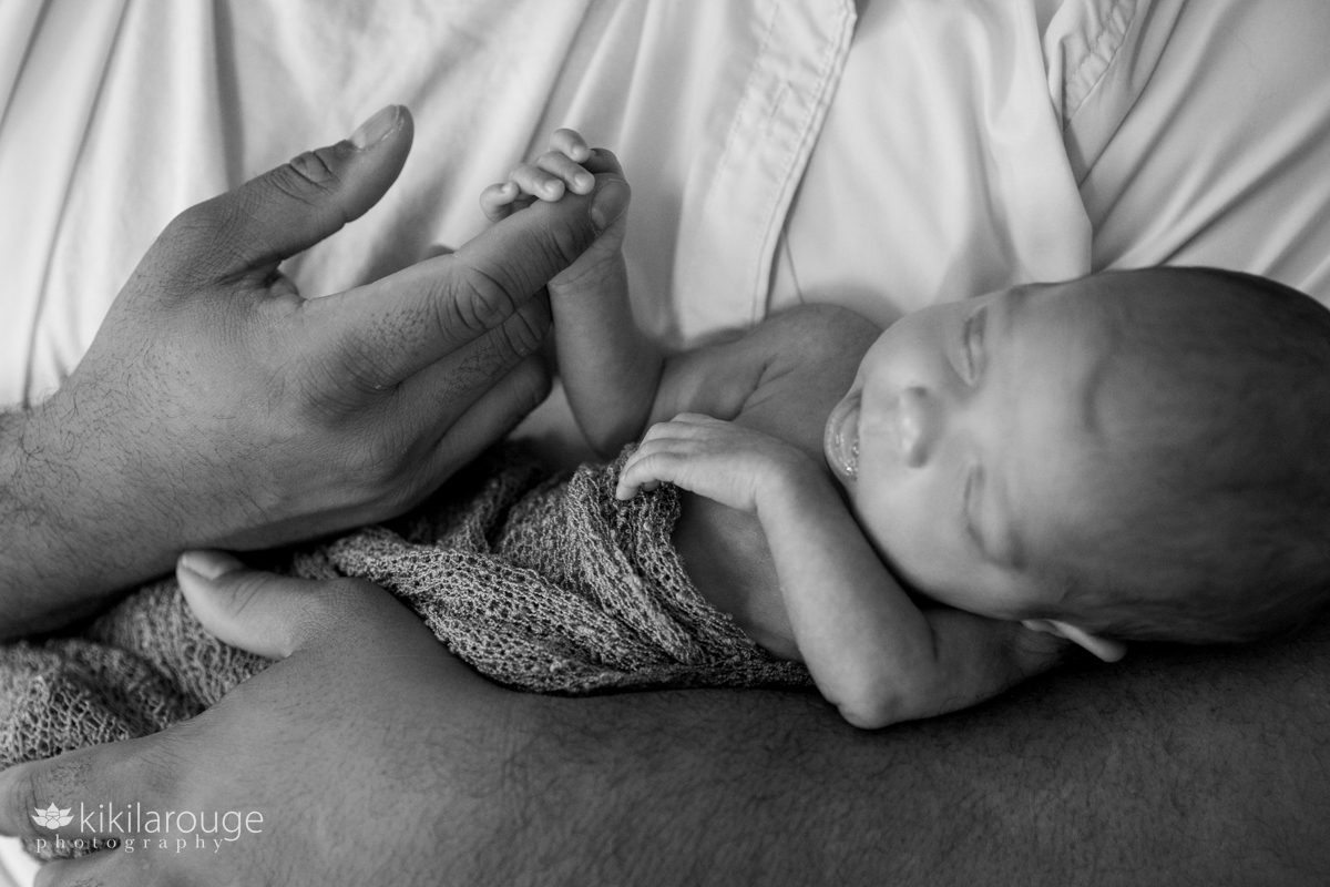 BW photo of newborn baby in father's arms holding on to his index finger with her tiny hand
