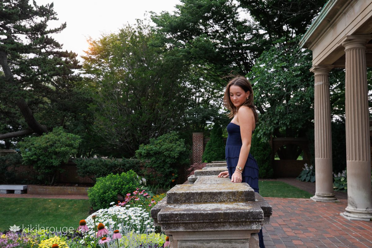 Teen girl leaning on stone wall in Rose Garden dressed in long navy sundress looking down