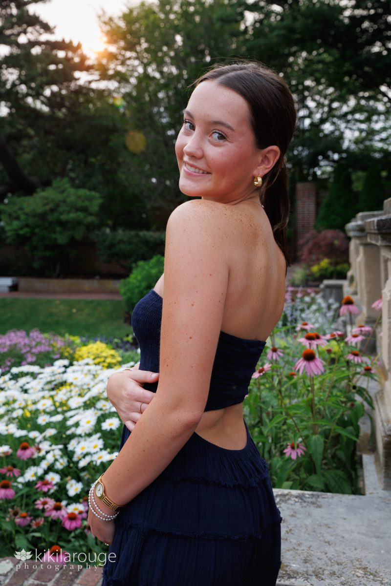 Senior portrait of teen brunette girl in blue sundress in front of flower garden with hazy sun flare