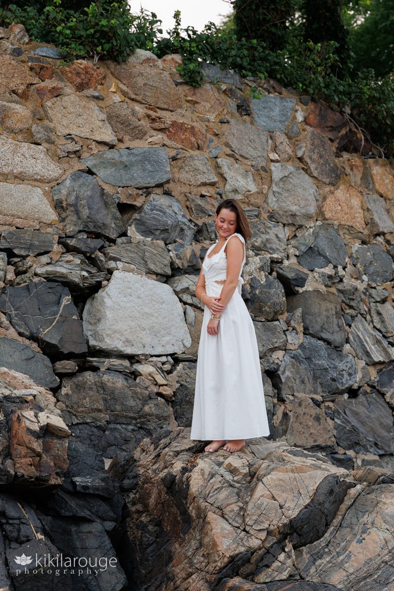 Senior girl in long white dress standing on rock wall holding her arm looking down smiling