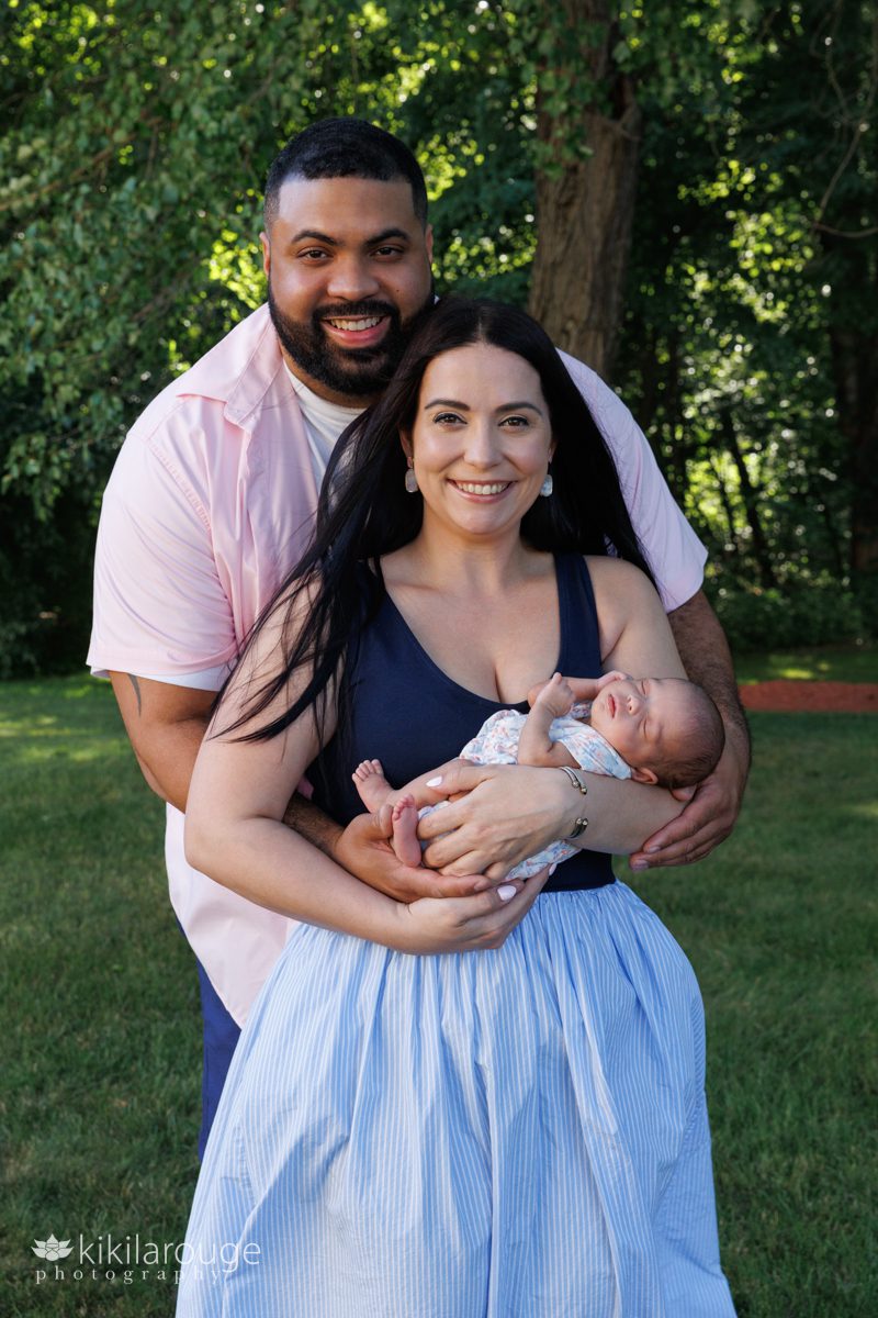 Couple in yard with trees in backdrop Mom in Blue dress and Dad in pink shirt smiling holding their newborn infant girl