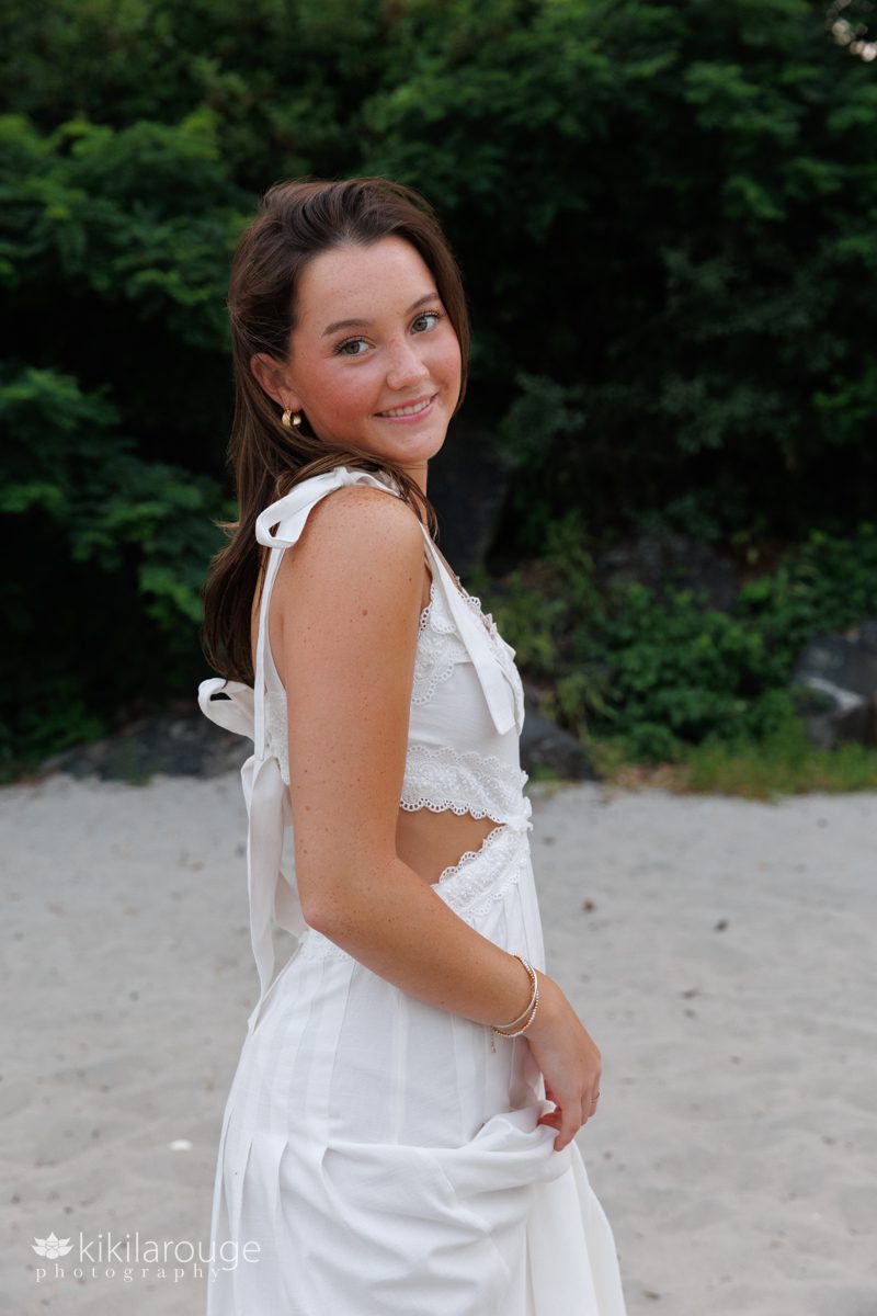 Teen Senior girl holding white dress with bows looking over shoulder smiling with dark green backdrop