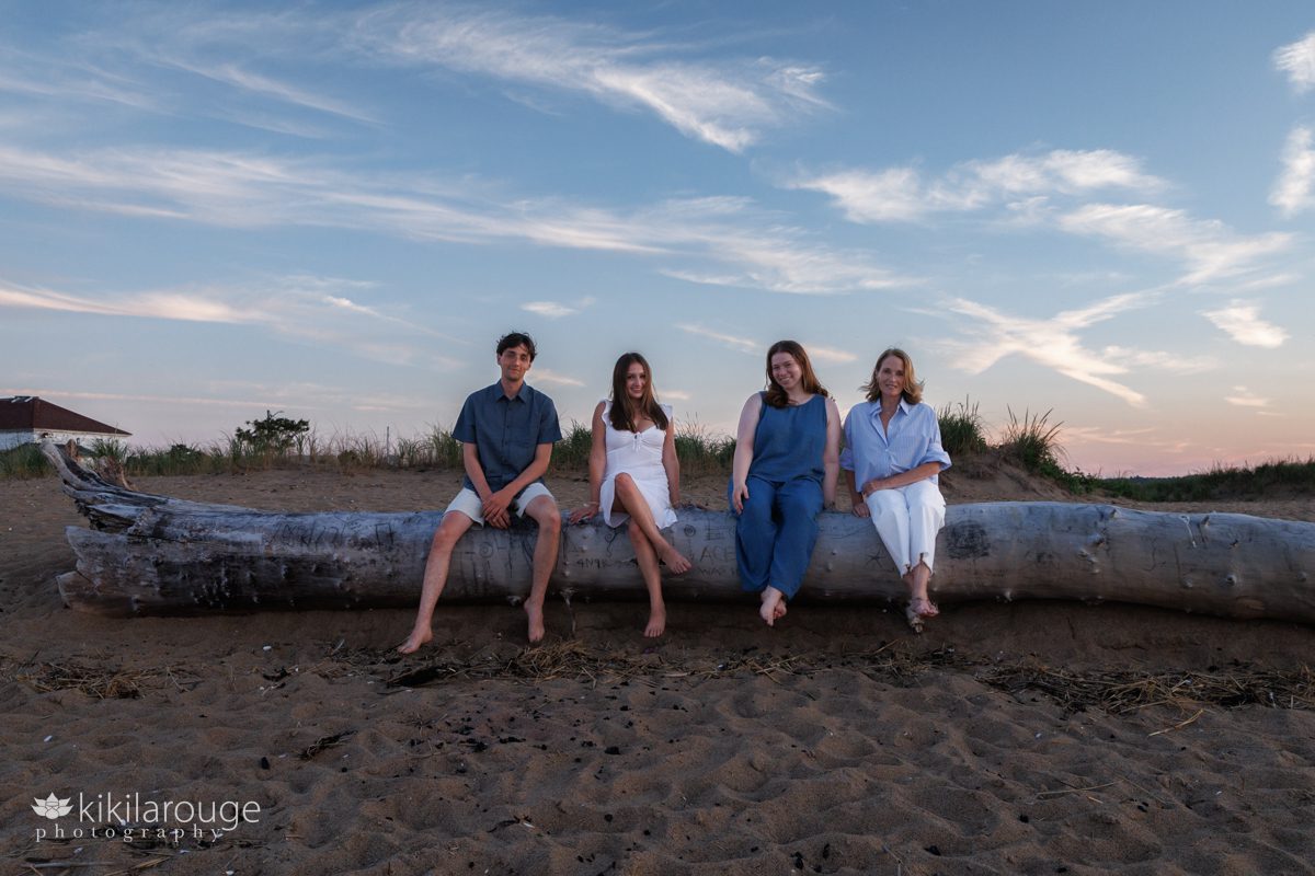Family of four sitting on large piece of driftwood with dramatic skies and old coast guard house in backdrop