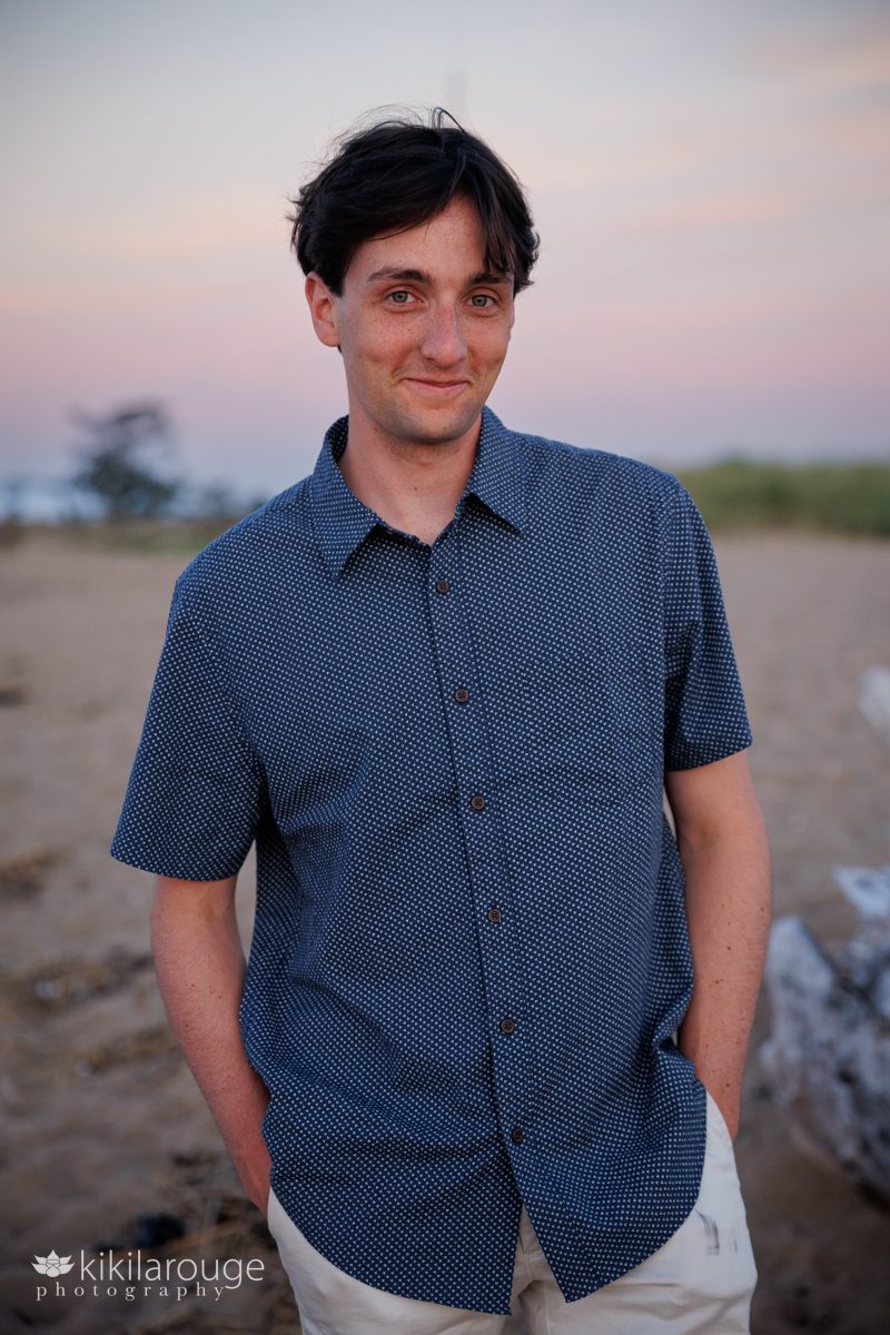 Handsome man with brunette hair and blue button up top with stars hands in short pockets smiling at the beach during sunset