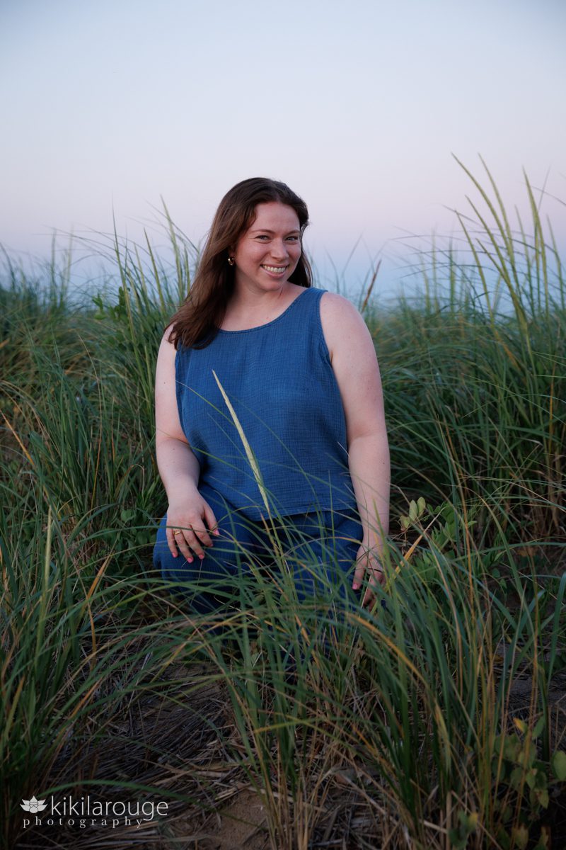 Woman with red hair and blue two pieces sleeveless summer comfy outfit sitting in beach dunes with pink sky backdrop